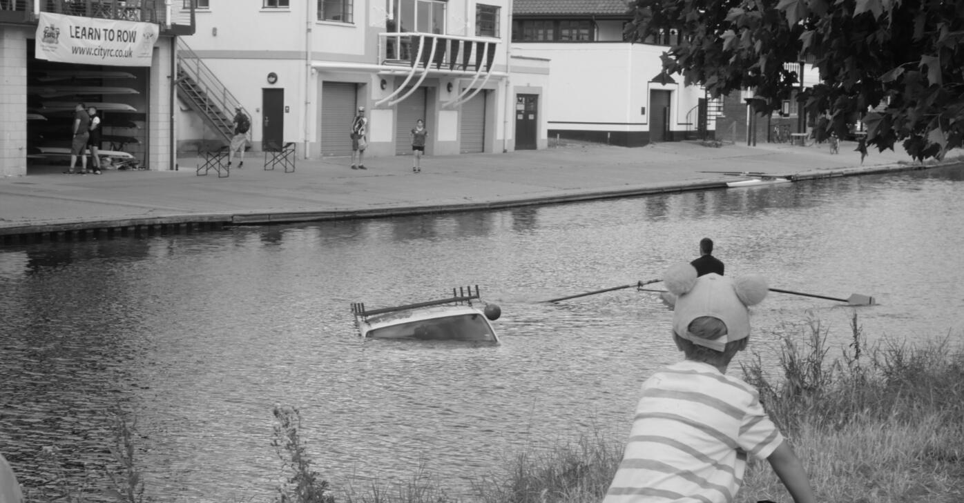 Boy cycles by the River Cam as a car sinks into the water and a rower sails past.